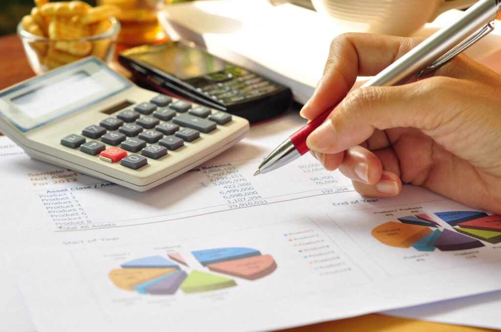 Eine Hand hält einen Stift über Finanzunterlagen mit Diagrammen; daneben liegen ein Taschenrechner und ein Handy.