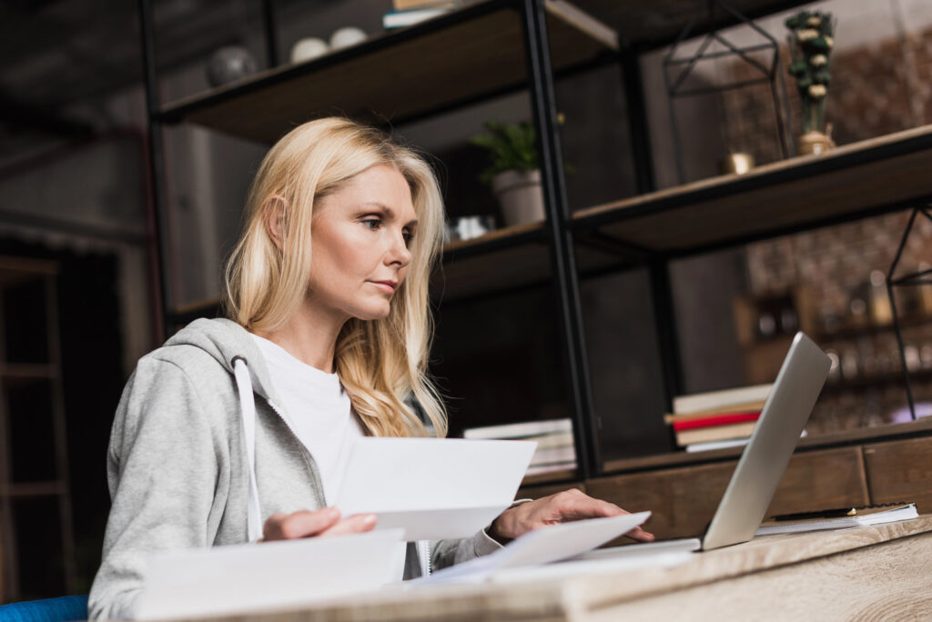 Junge Frau sitzt am Schreibtisch vor einem Laptop und sortiert Unterlagen in einem modernen Büro.