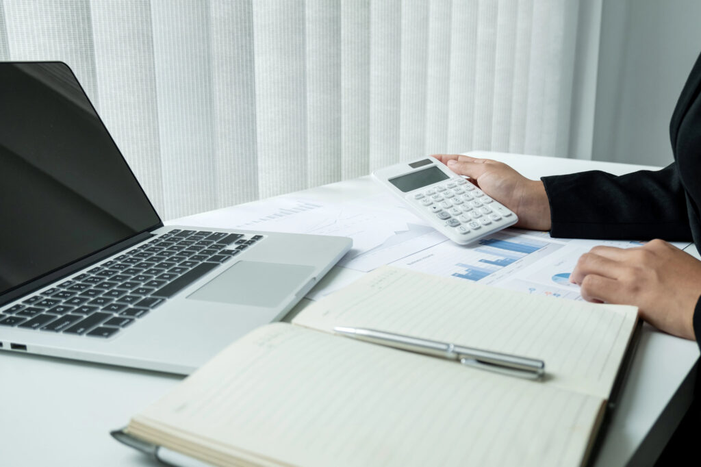 Person arbeitet am Schreibtisch mit Laptop, Taschenrechner und Finanzunterlagen mit Diagrammen.